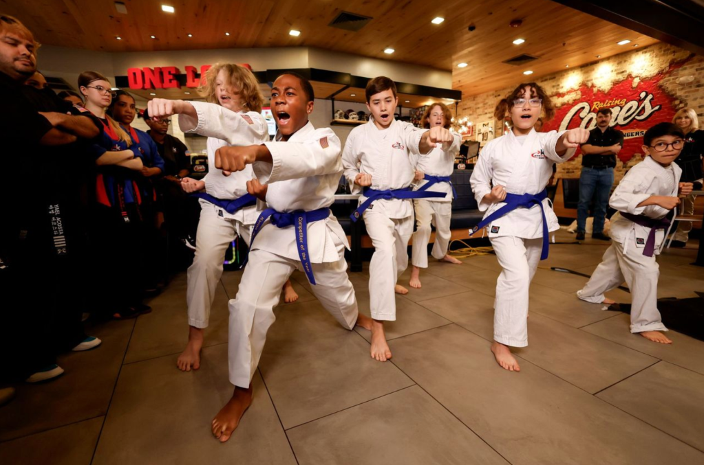 A group of children in white martial arts uniforms performing karate moves in a martial arts studio with a "Raising Cane's" logo visible.