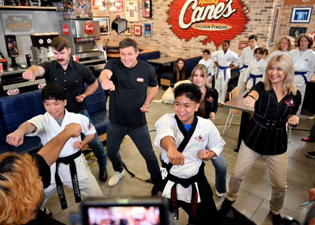 A group of people in martial arts uniforms practicing karate moves in a Raising Cane's restaurant, with the Raising Cane's logo prominently displayed on the wall.