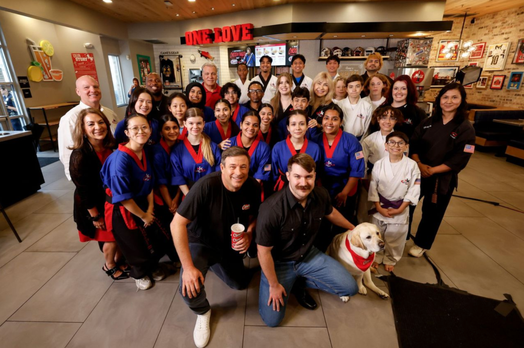 A large group of people, including adults and children, posing together in a restaurant or cafe setting. The group is wearing uniforms or matching outfits, and there is a dog present.