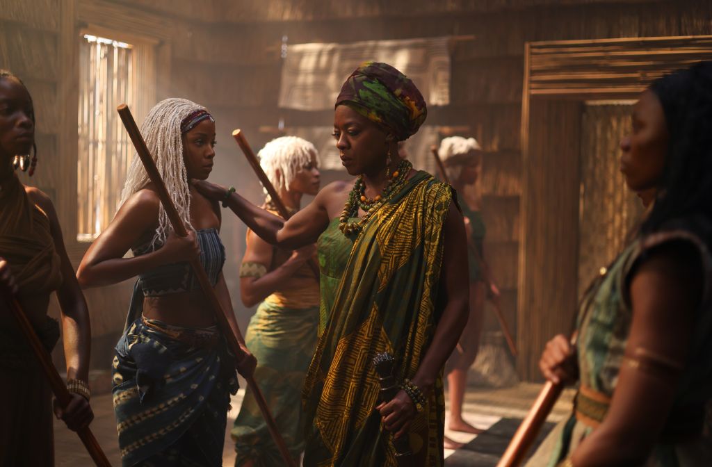 A group of people in traditional African attire, including a woman in a yellow and black patterned dress and headwrap, interacting in an indoor setting.