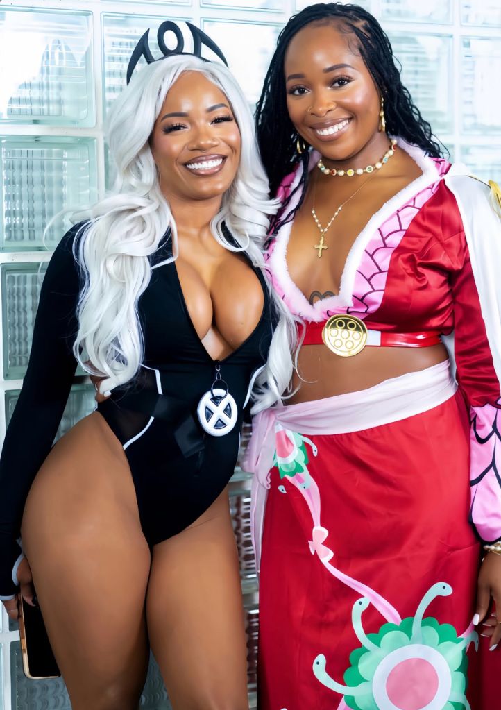 Two smiling women in colorful superhero-inspired costumes posing together in front of a window.