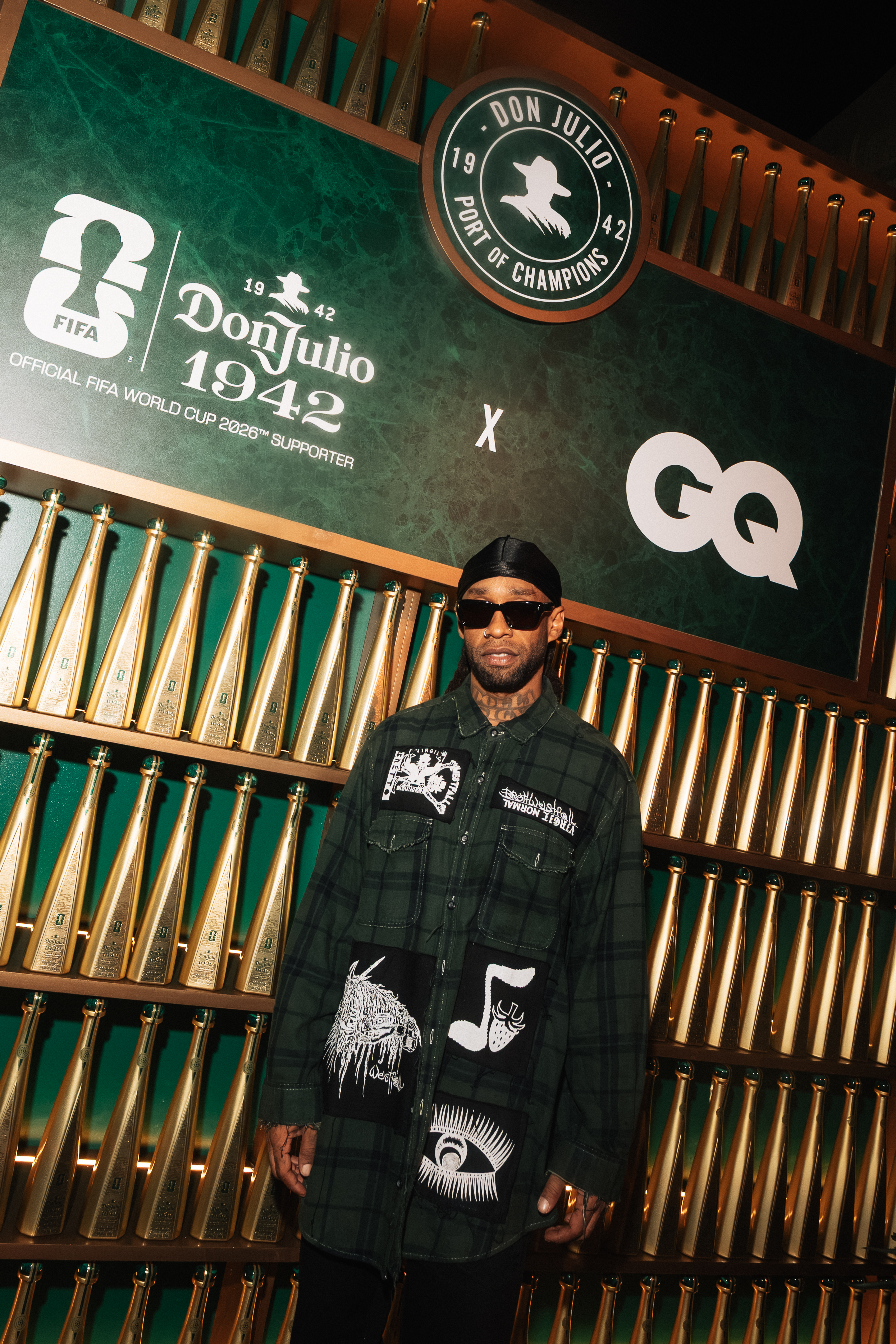 A person wearing a green and black plaid jacket and sunglasses stands in front of a wall displaying the "Don Julio 1942" logo and "GQ" branding.