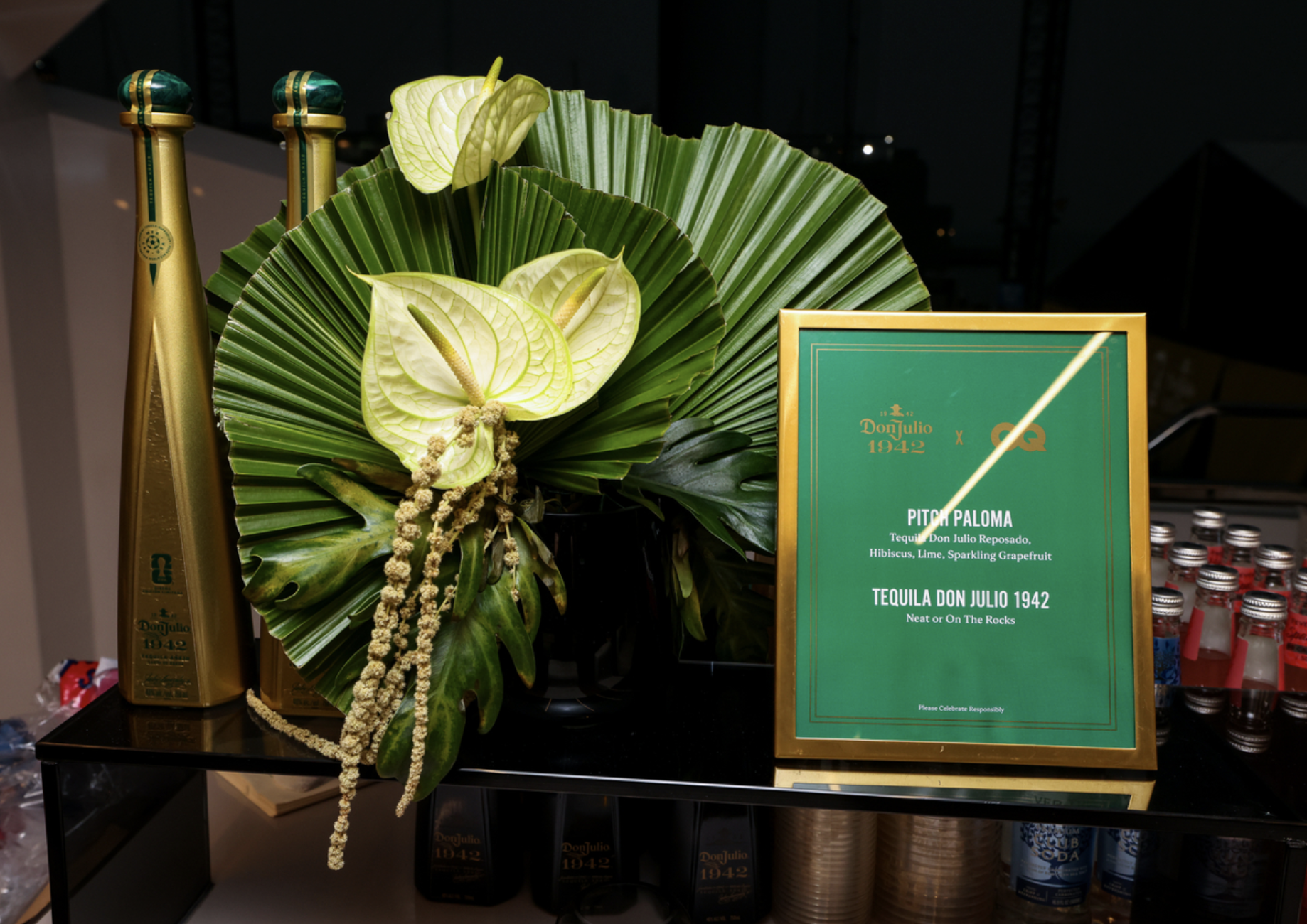 A display of tropical flowers and leaves, including anthurium and palm fronds, alongside bottles of Tequila Don Julio 1942 and a framed advertisement for the "Pitch Paloma" cocktail.