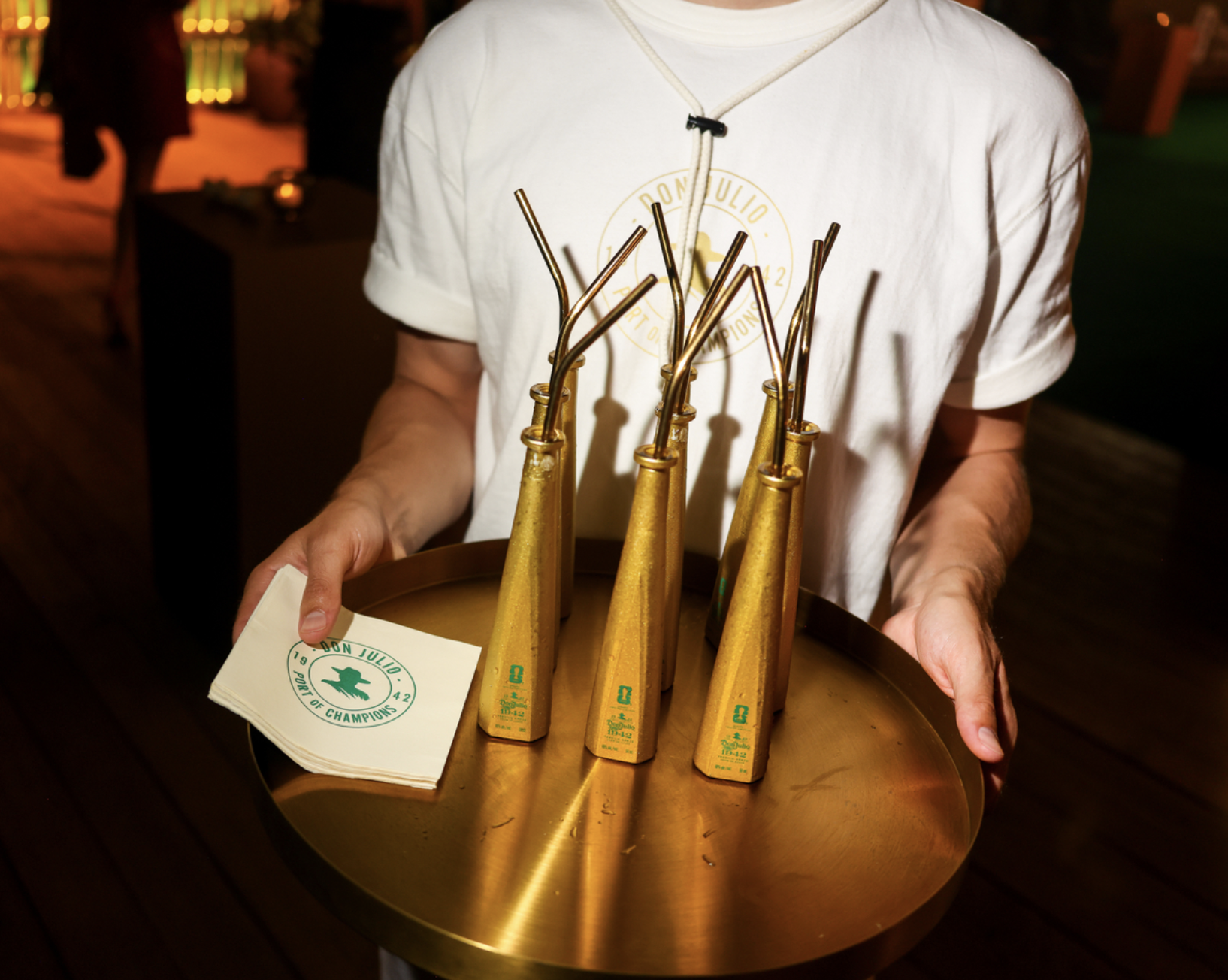 A person holding a tray with three golden trophies or awards, wearing a white t-shirt with the text "Corn Toss Champions".