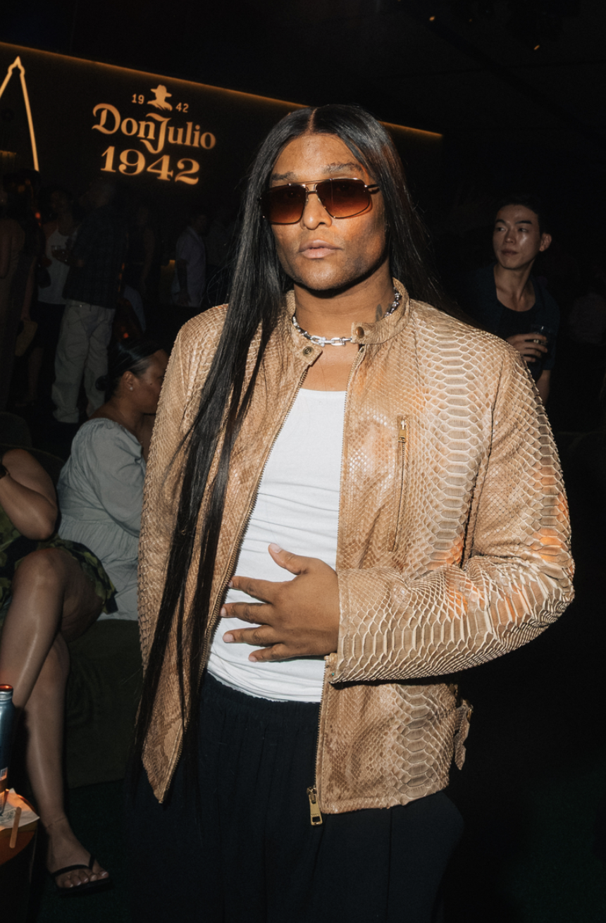 A person wearing a beige snakeskin-patterned jacket and sunglasses stands in front of a sign that says "Don Julio 1942".