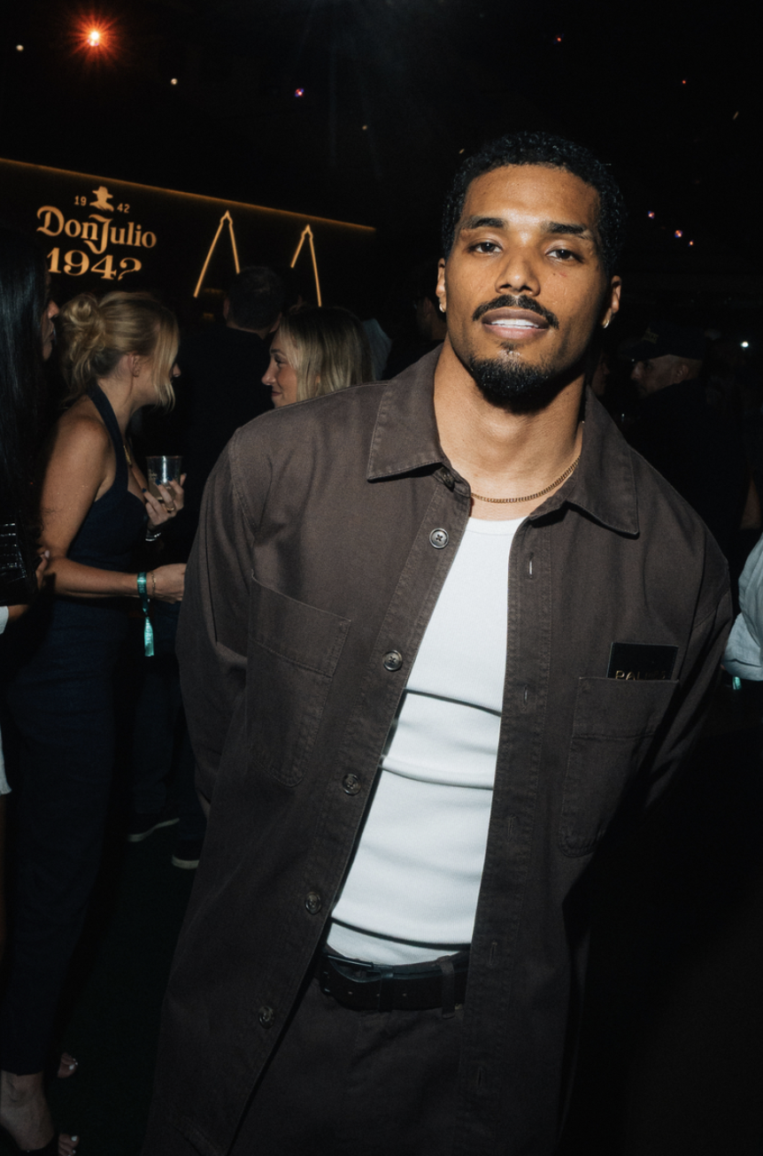 A man with a beard wearing a dark jacket stands in front of a sign that says "Don Julio 1942" at a nighttime event.