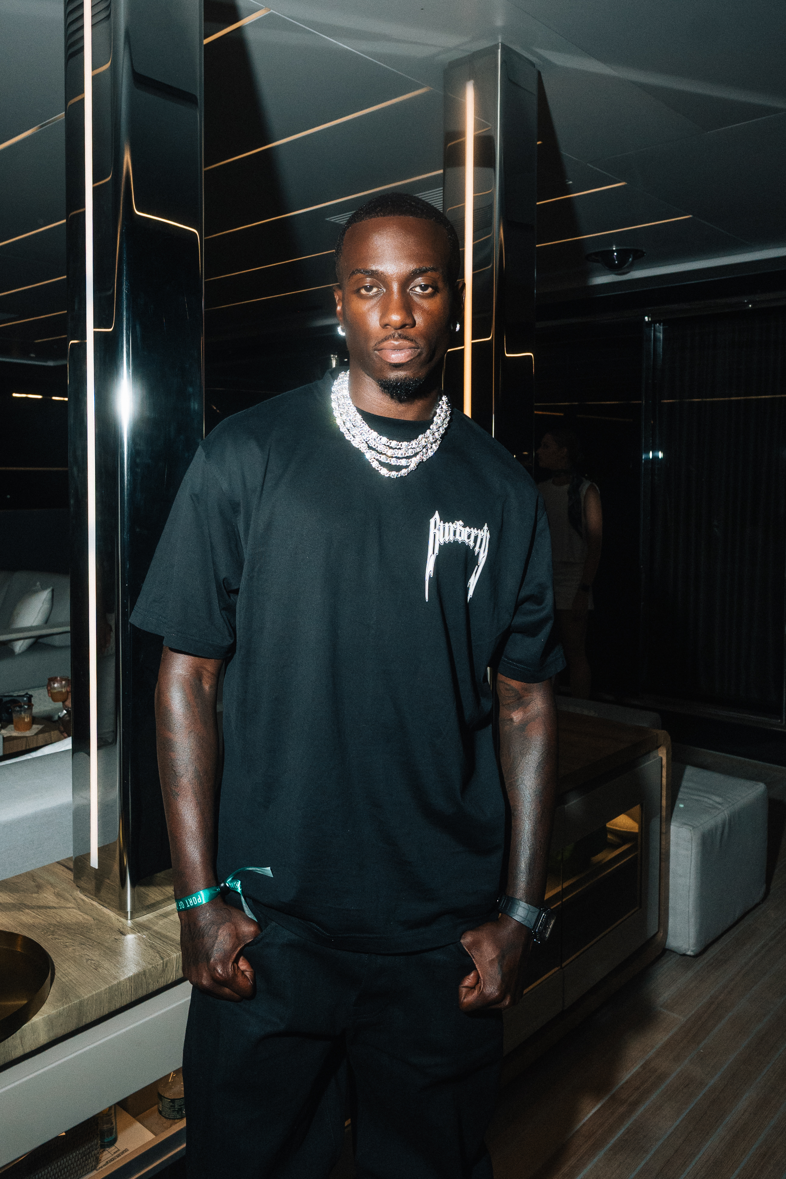 A young Black man wearing a black t-shirt with "Burberry" text and multiple diamond necklaces, standing in a dimly lit room with geometric patterns on the walls.