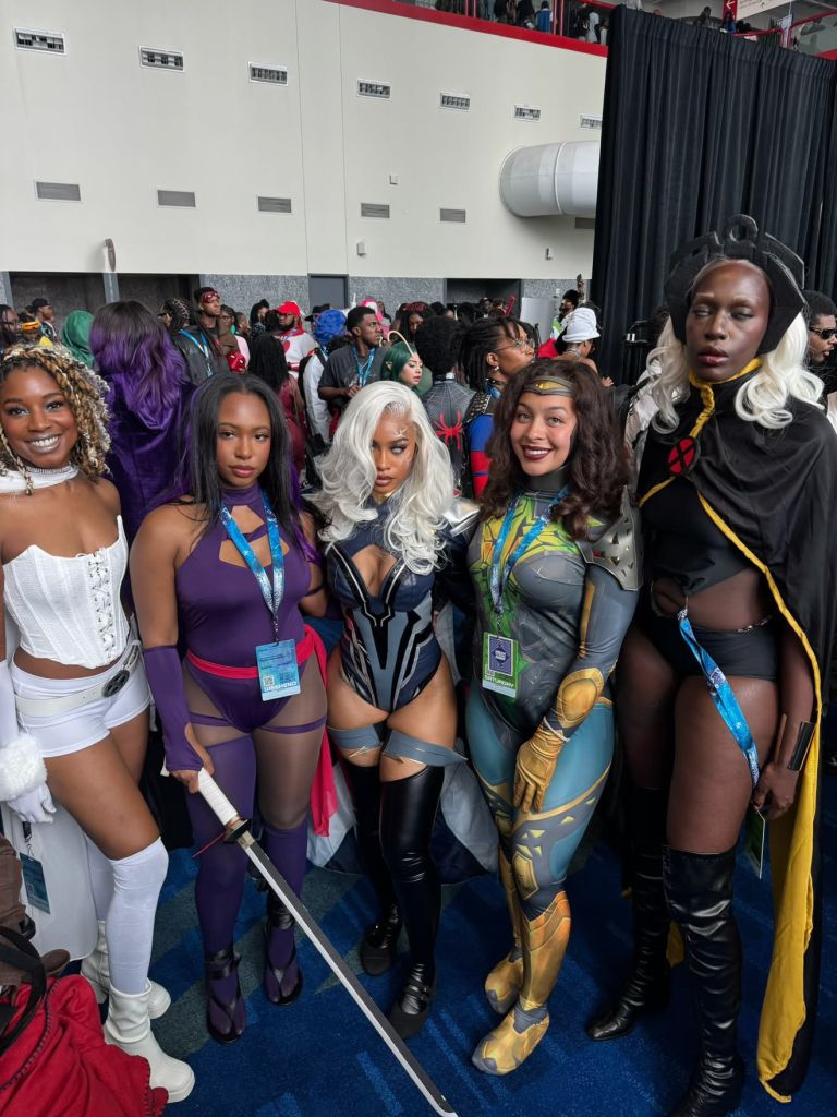 A group of people dressed in colorful, elaborate costumes and superhero-inspired outfits posing together in a convention center hallway.
