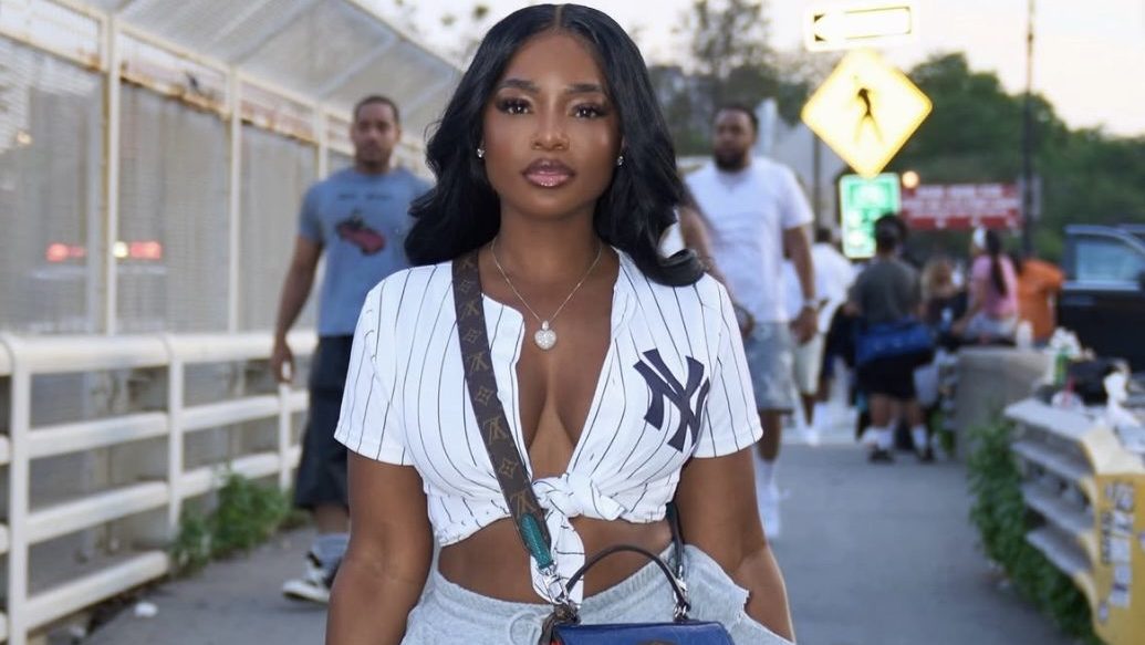 A woman wearing a white pinstriped NY Yankees jersey and holding a blue handbag stands in an urban setting with other people in the background.