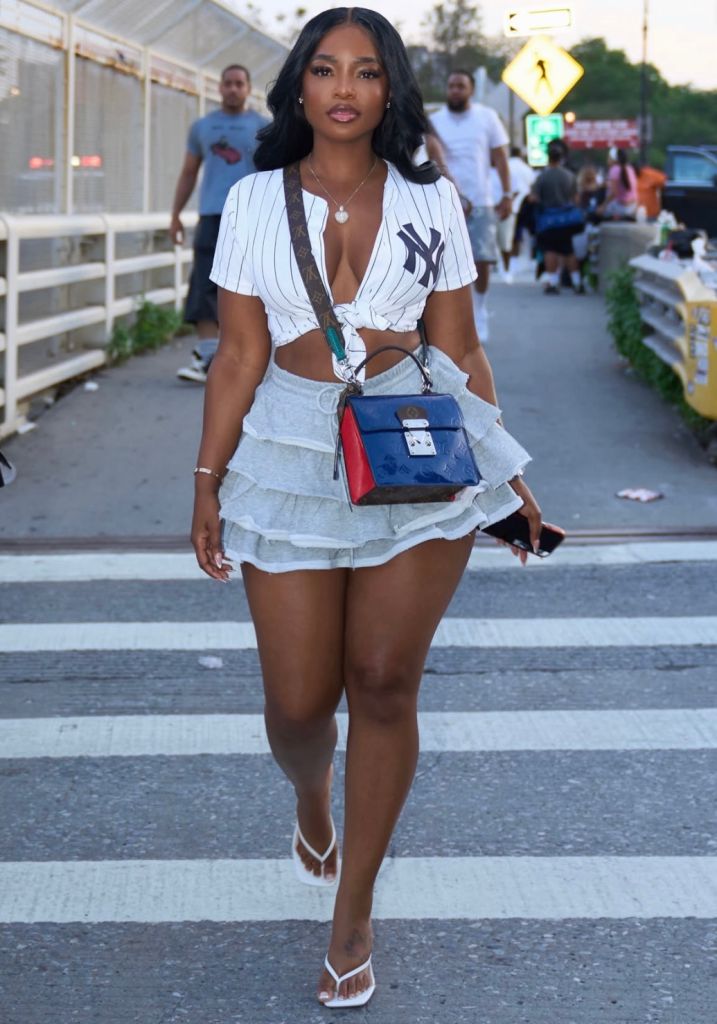 A woman wearing a white pinstriped Yankees jersey and a short skirt, carrying a blue handbag, walking on a city street.