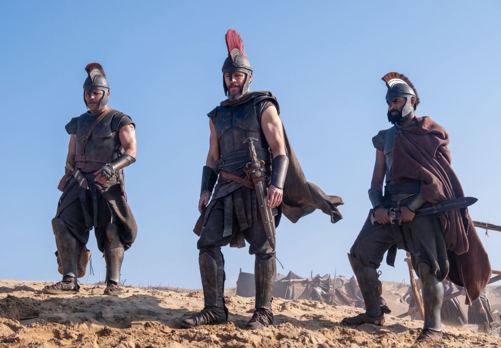 Three armored warriors in ancient Greek-style uniforms standing in a desert landscape.
