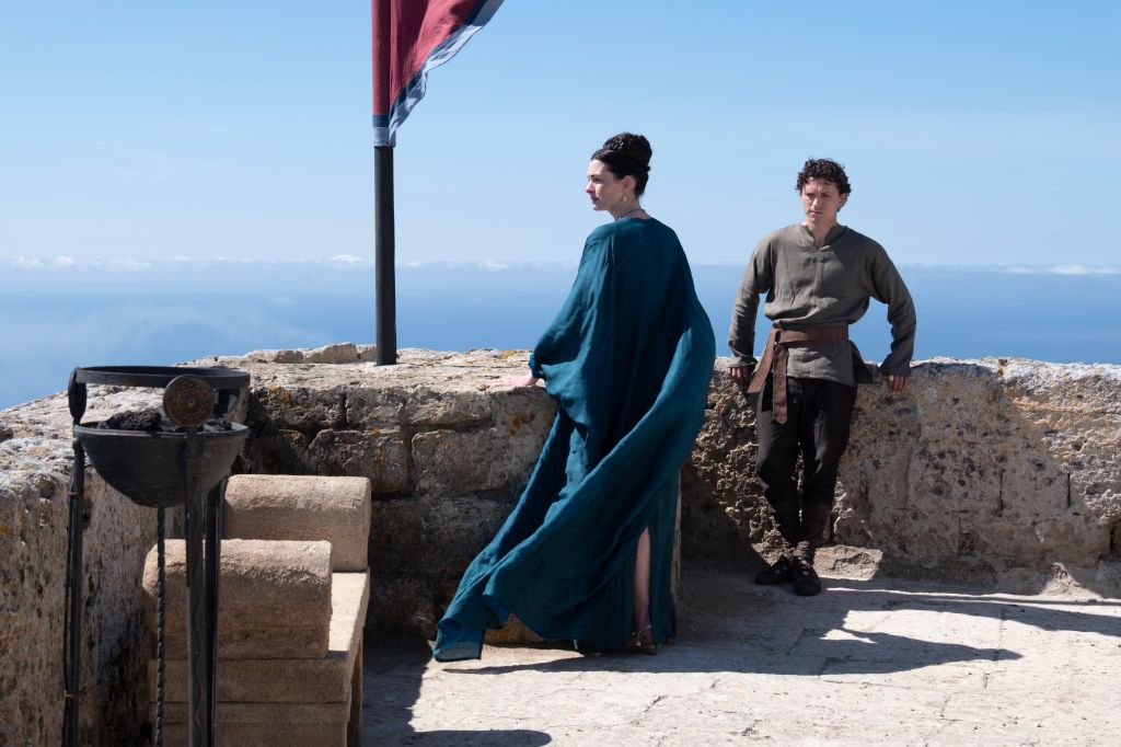 Two people standing on a rocky outcrop overlooking a body of water, with a red flag waving in the background.