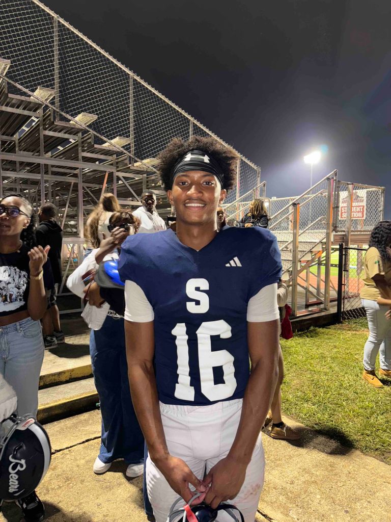 A smiling young person wearing a navy blue jersey with the number"516" standing on a sports field at night.
