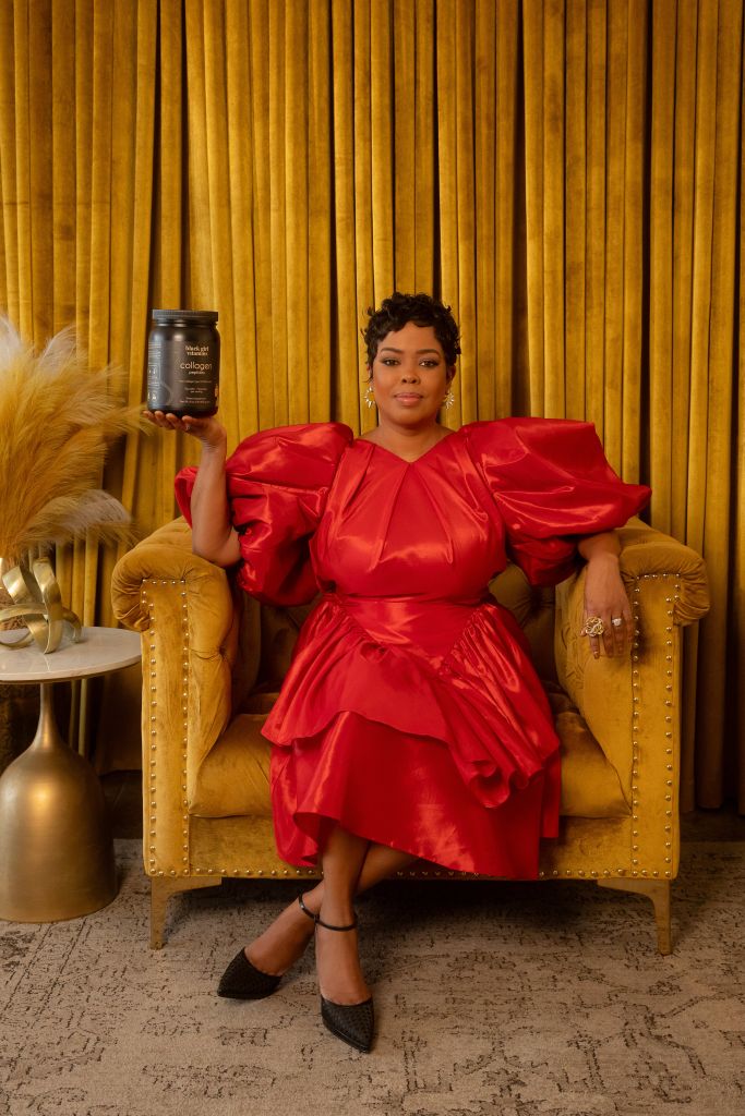 A woman in a red puff-sleeved dress holds a collagen supplement bottle, seated in a gold-accented chair against a curtained backdrop.