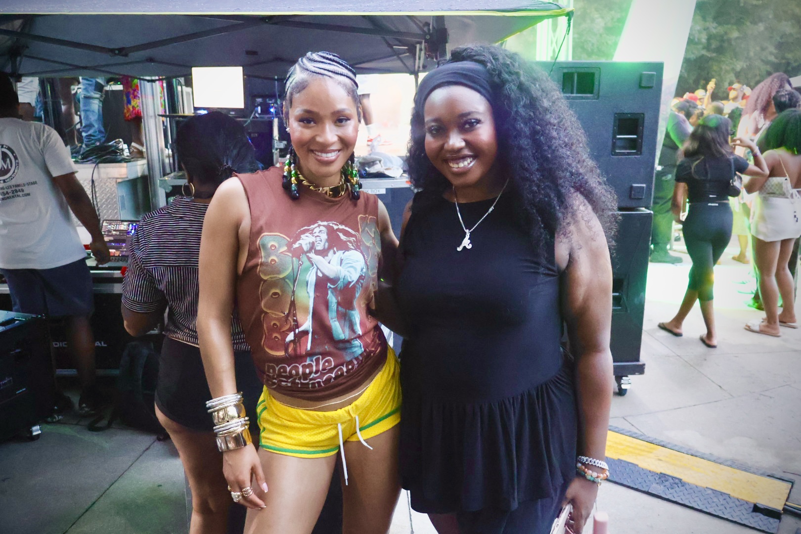 Two smiling women standing in front of a green backdrop, one wearing a colorful graphic tank top and the other wearing a black dress.