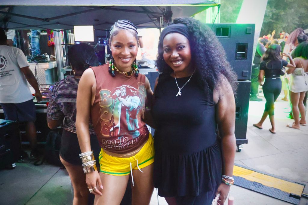 Two smiling women standing in front of a green backdrop, one wearing a colorful graphic tank top and the other wearing a black dress.