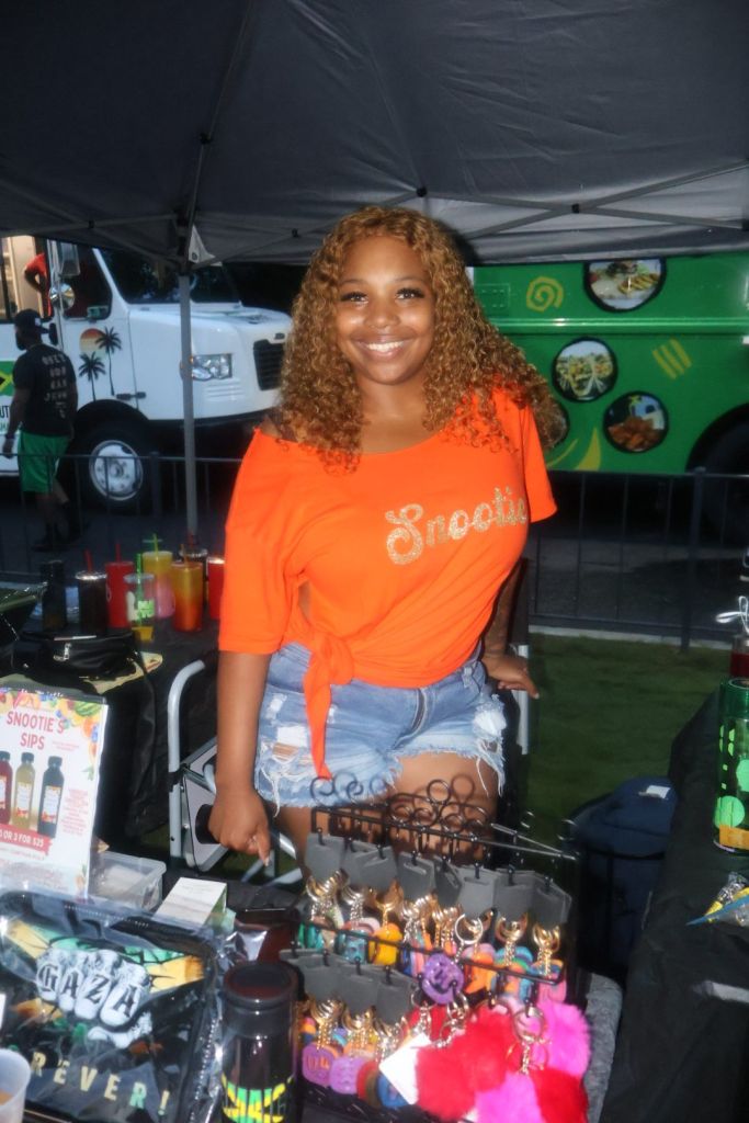 A smiling woman with curly hair wearing an orange shirt with "Snootie" text, standing in front of a green food truck at an outdoor event.
