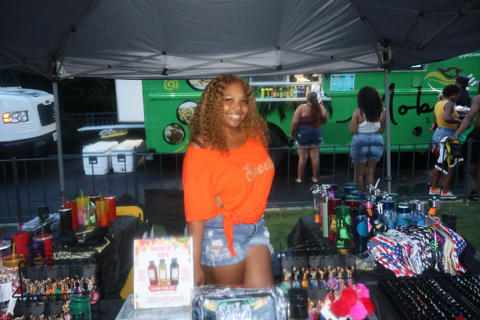 A smiling woman with curly hair stands in front of a green food truck, surrounded by various merchandise and supplies.