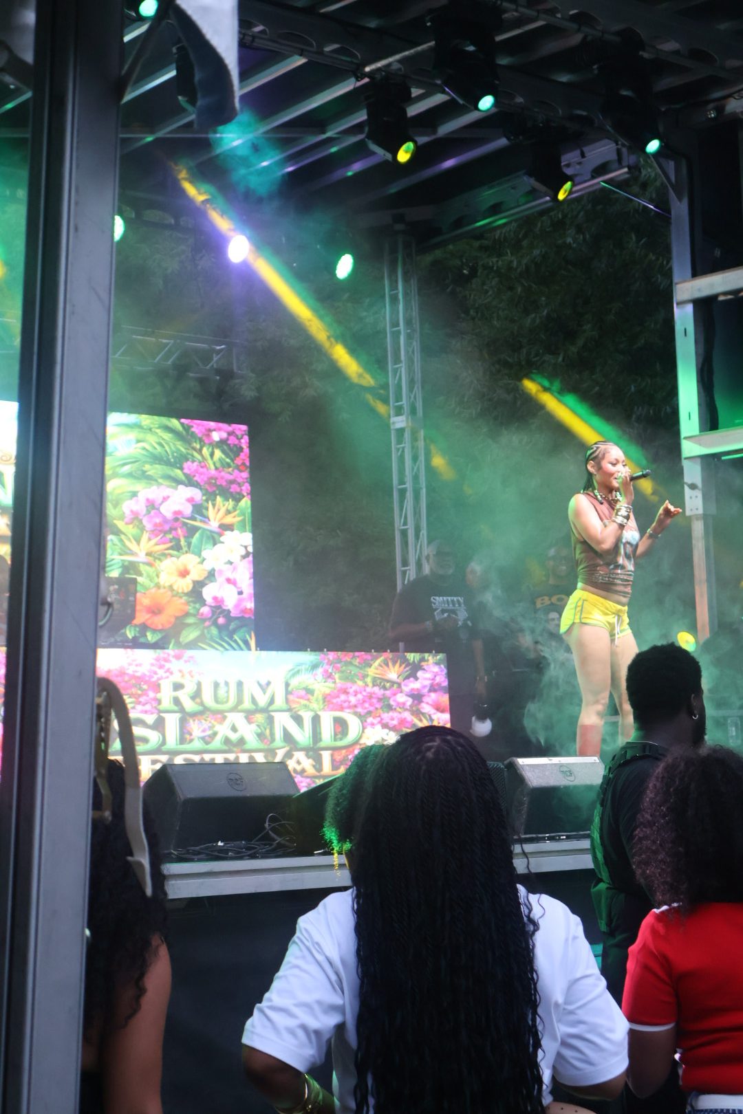 A performer on stage at the Rum Island Festival, singing into a microphone while surrounded by colorful lighting and a large floral backdrop.