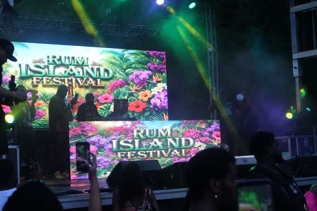 Rum Island Festival event with colorful tropical floral backdrop and silhouetted crowd in front of stage.