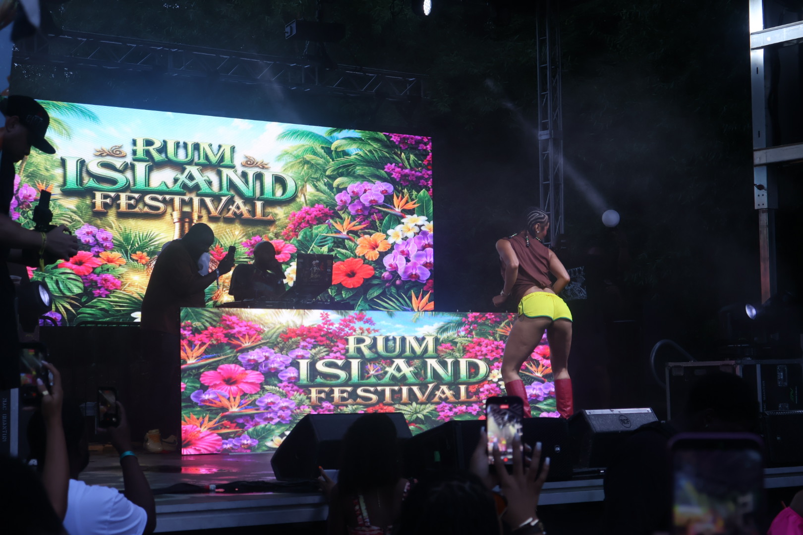 Rum Island Festival event with tropical floral backdrop and silhouetted figures on stage.
