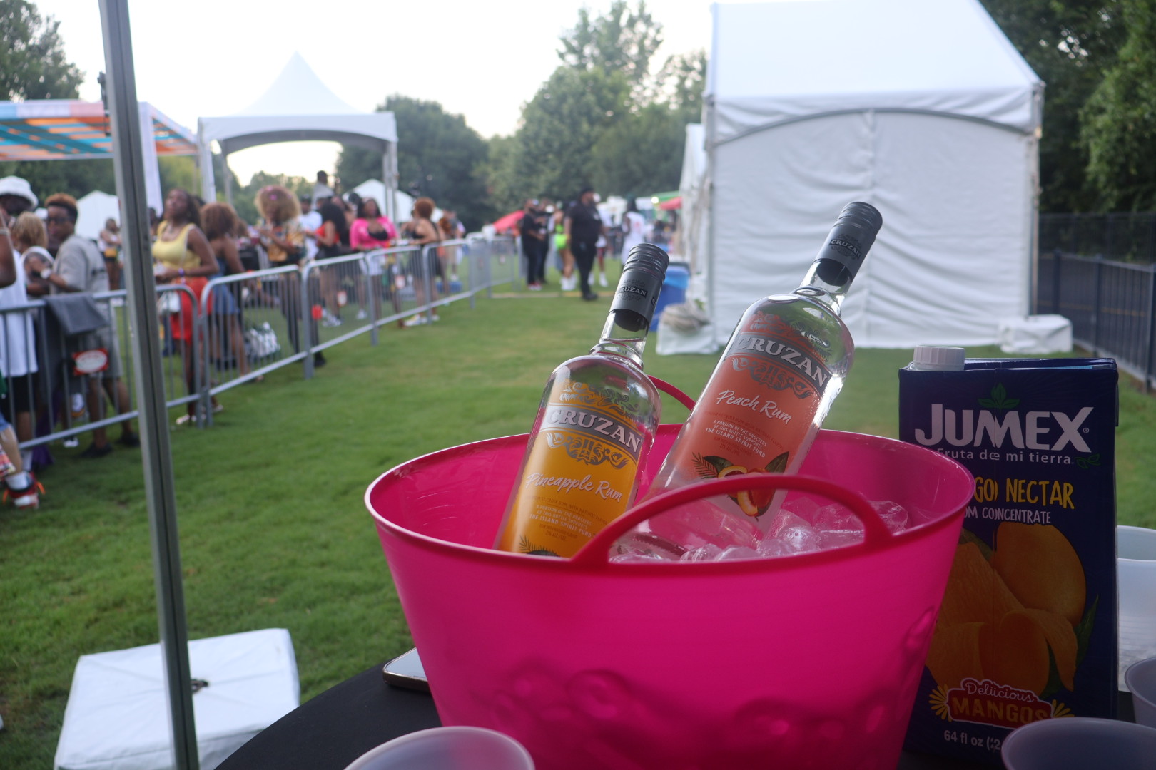 Outdoor festival with a table displaying bottles of Cruzan rum and Jumex fruit nectar concentrate in a large pink bucket filled with ice.