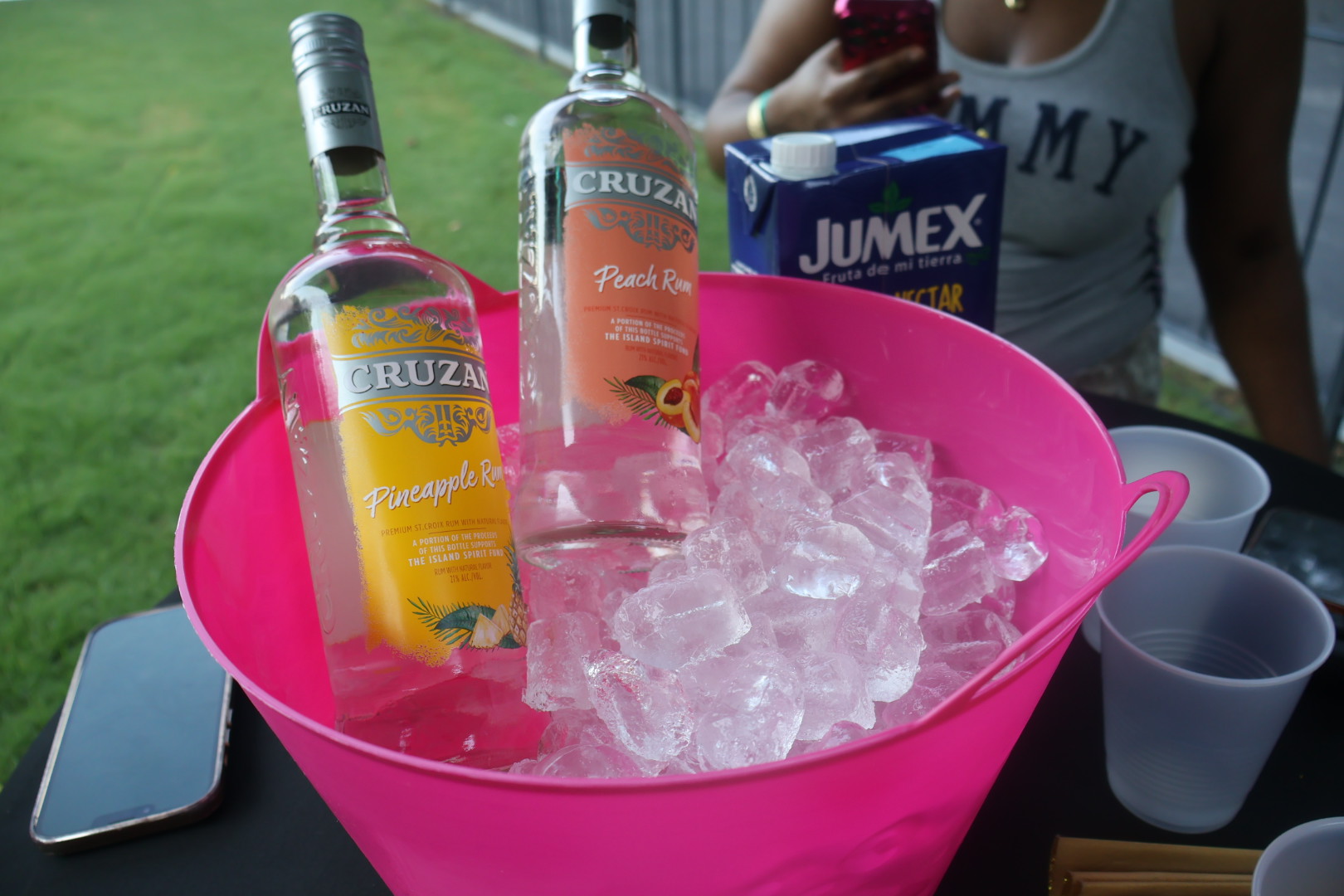 A pink plastic tub filled with ice cubes, bottles of Cruzan rum, and a carton of Jumex juice on a grassy surface.