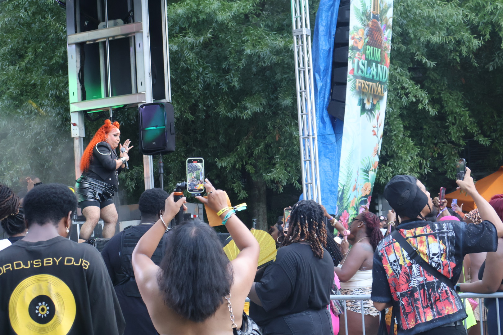 A person with bright orange hair performing on a stage at an outdoor festival, surrounded by a crowd taking photos with their phones.