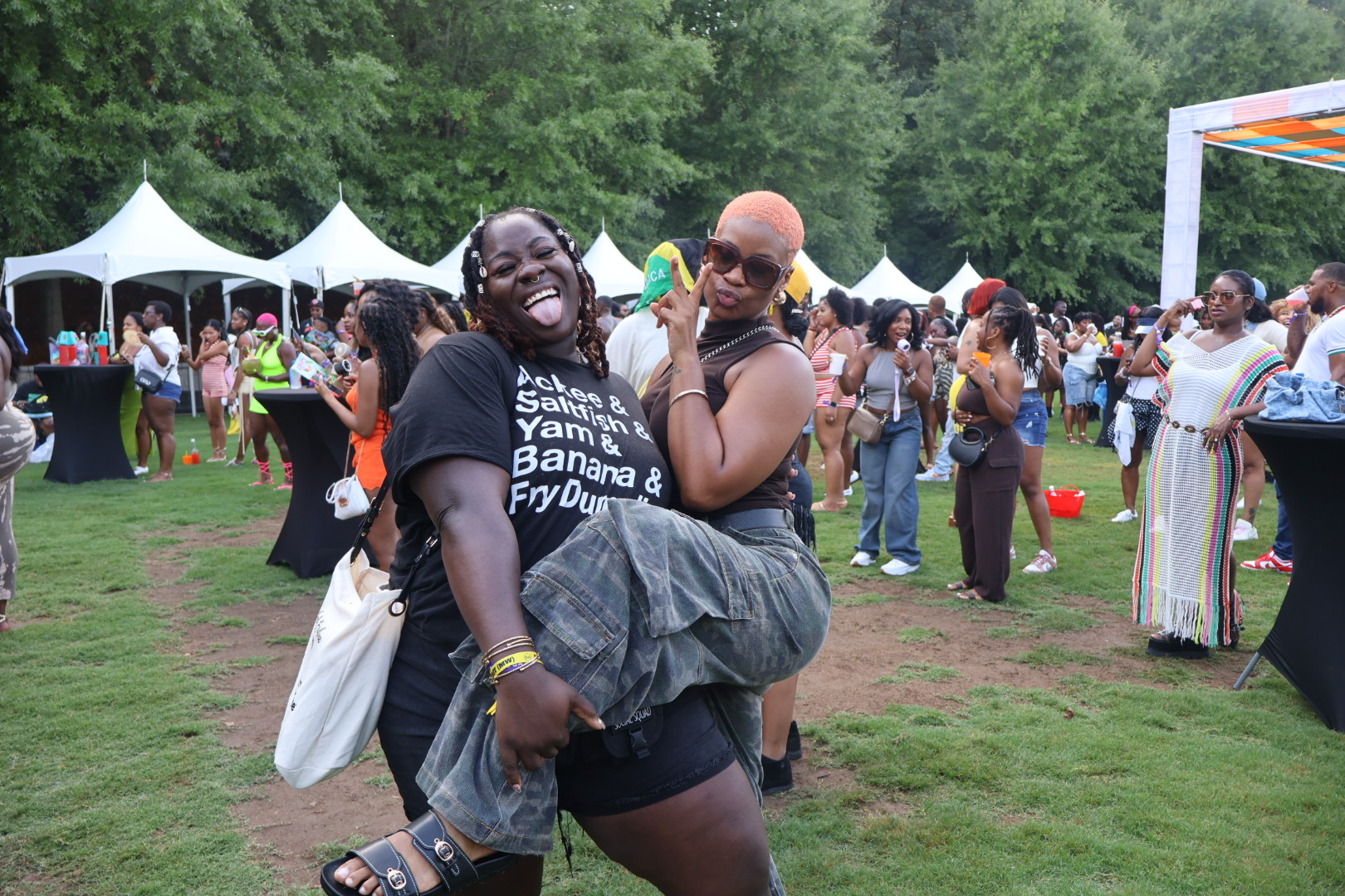 A group of people enjoying an outdoor festival, with tents and colorful decorations in the background. A person wearing a shirt that says "Banana & Fry Dun" is laughing and embracing another person.