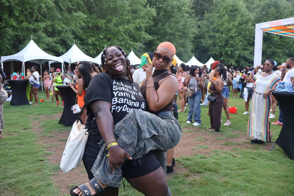 A group of people enjoying an outdoor festival, with tents and colorful decorations in the background. A person wearing a shirt that says "Banana & Fry Dun" is laughing and embracing another person.
