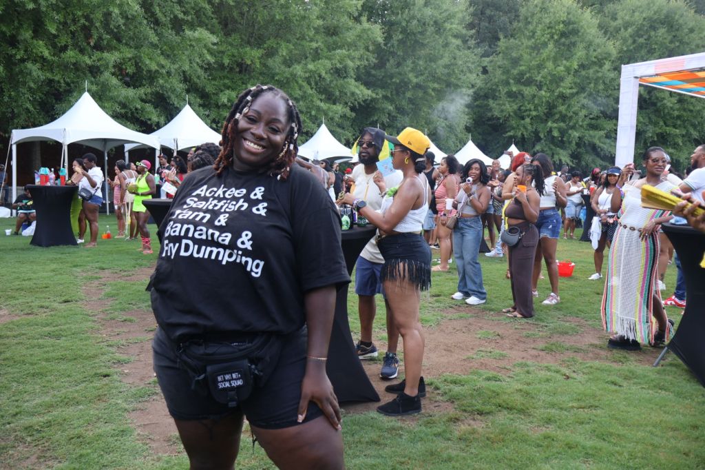 A smiling person wearing a t-shirt with the text "Ackee & saltfish & yam & banana & my Dumping" standing in a crowd at an outdoor event.