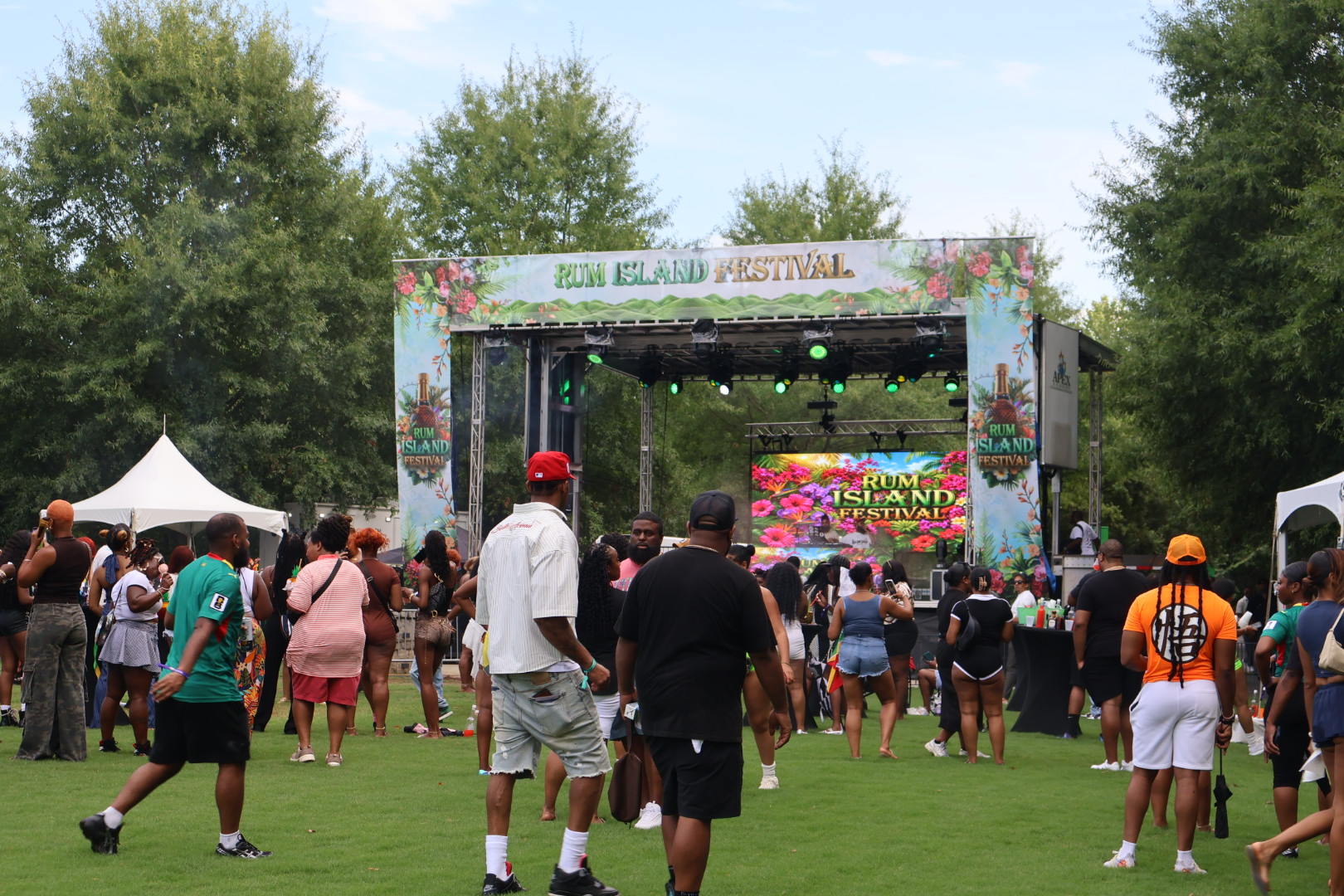 Outdoor music festival with a large stage, crowd of people, and colorful decorations reading "Rum Island Festival".