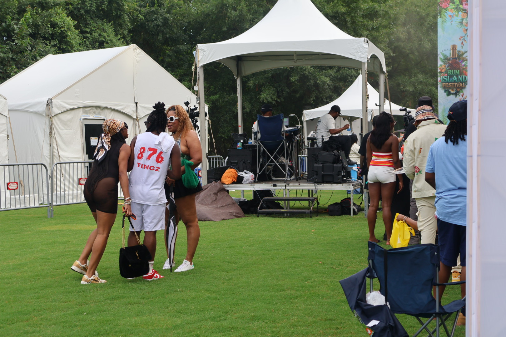 Outdoor festival with tents, people in casual summer attire, and a stage with audio equipment.