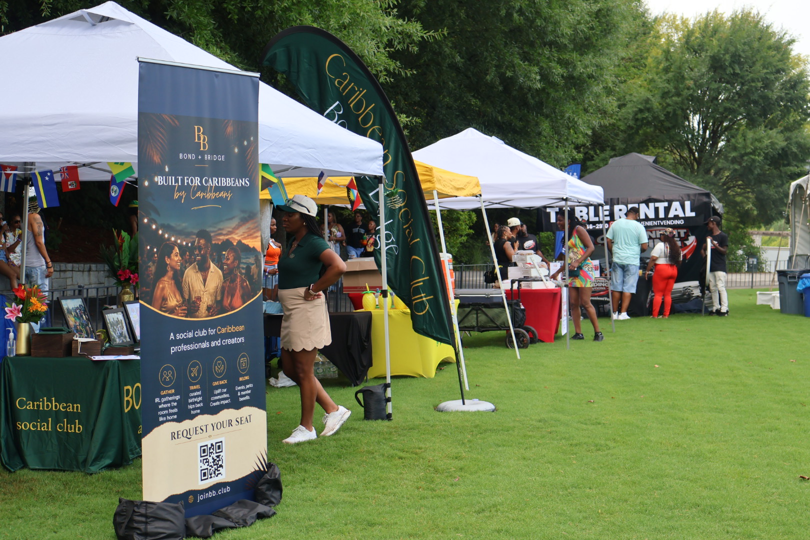 Outdoor event with Caribbean social club booth, tents, and people browsing merchandise and activities on a grassy field.