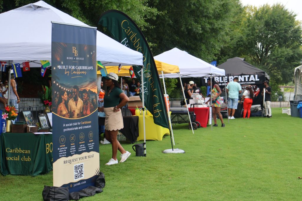 Outdoor event with Caribbean social club booth, tents, and people browsing merchandise and activities on a grassy field.