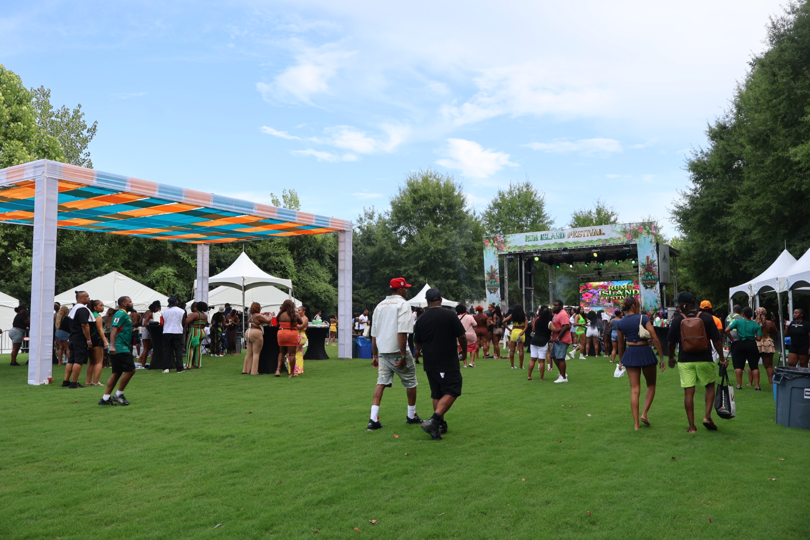 Outdoor festival with colorful tents, stage, and crowd of people enjoying the event on a grassy field surrounded by trees.