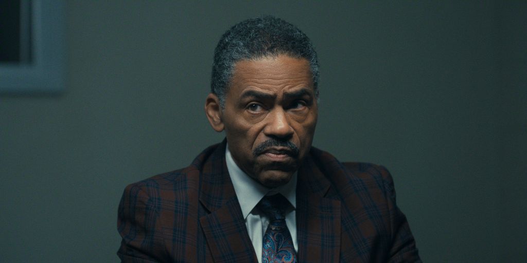 A serious-looking middle-aged Black man in a plaid suit and tie, with a stern expression on his face.