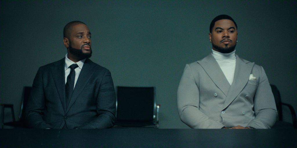 Two well-dressed Black men sitting in a dark room, one in a black suit and the other in a gray double-breasted suit.