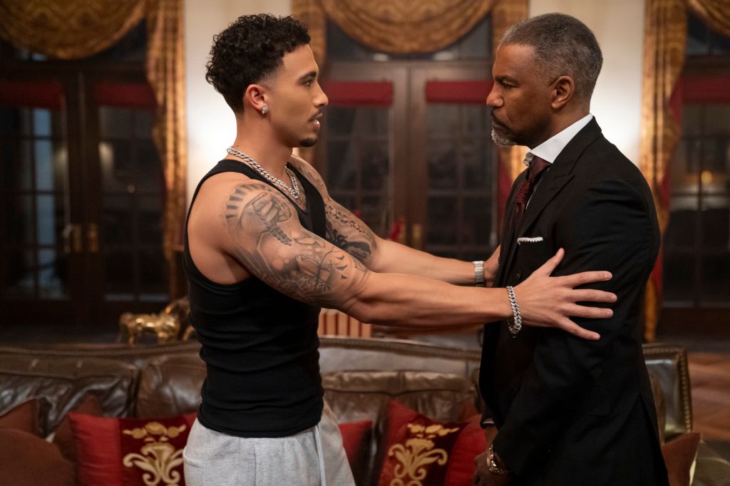 Two men, one in a black tank top with tattoos and the other in a suit, stand facing each other in a lavish room.
