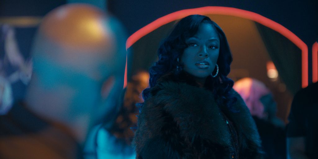 A woman with long dark hair wearing a fur-trimmed black coat stands in a dimly lit setting with colorful lights.