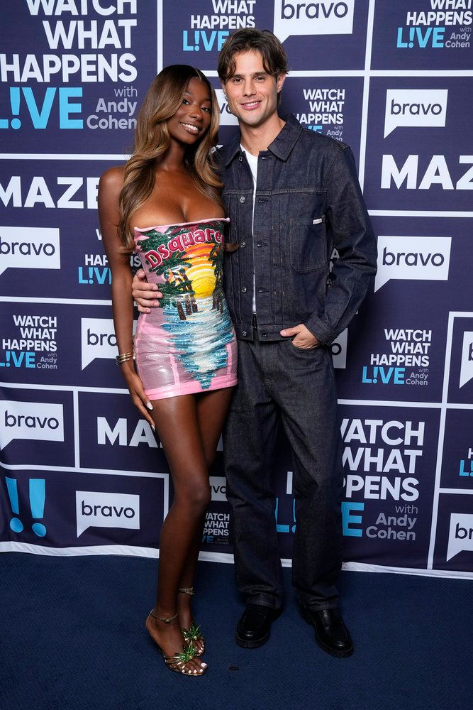Two people posing in front of a "Watch What Happens Live with Andy Cohen" backdrop, one wearing a colorful patterned dress and the other in a denim jacket.