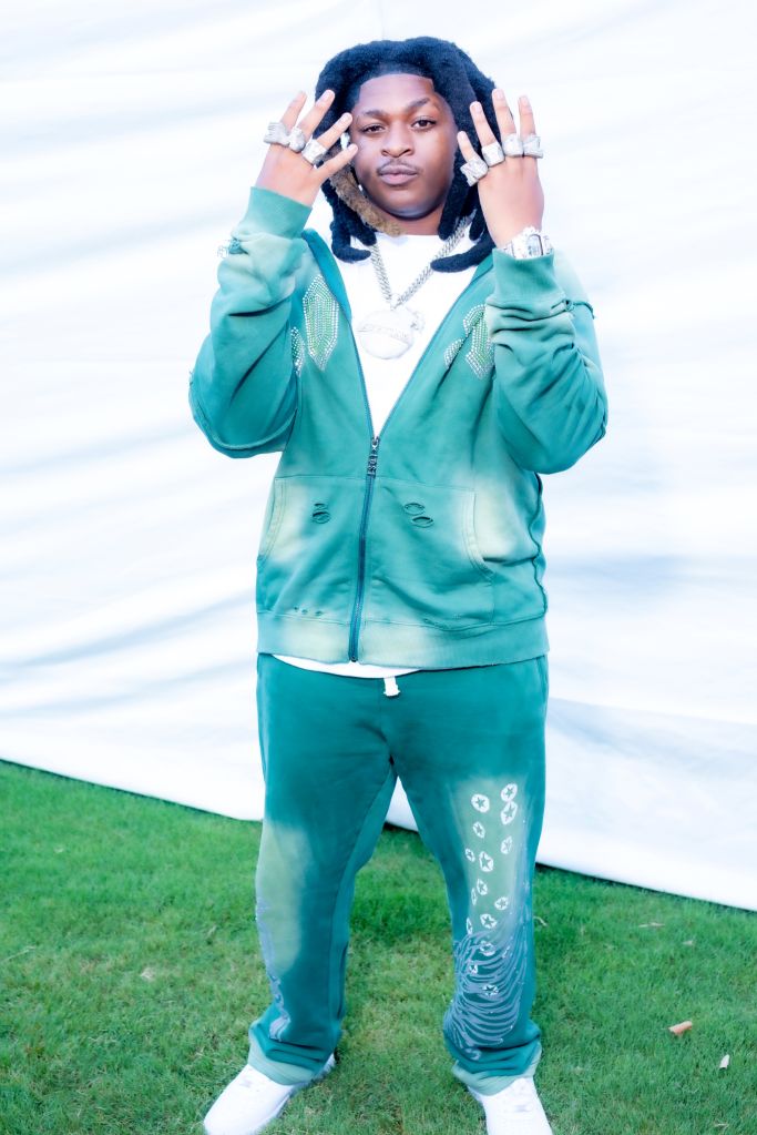 A person wearing a teal sweatsuit and jewelry stands with hands raised in a gesture, against a backdrop of grass and sky.