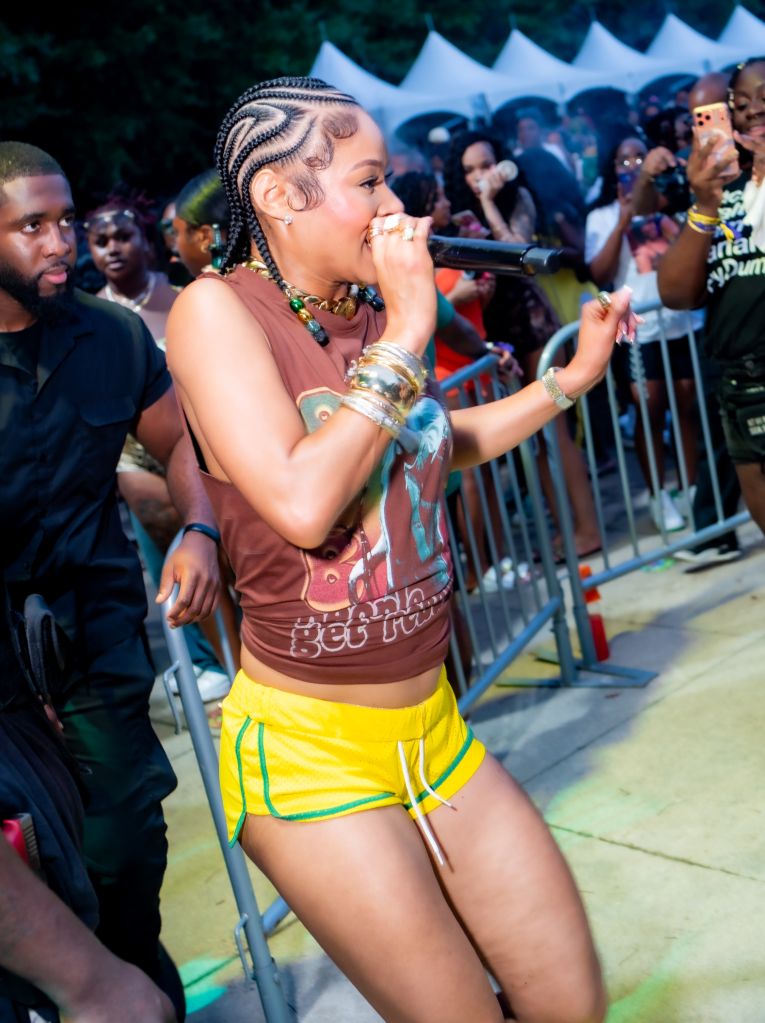 A woman in a pink top and yellow shorts performing on stage at a crowded outdoor event, holding a microphone and surrounded by spectators.