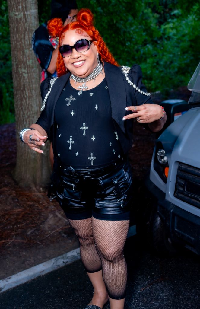 A smiling woman with bright red hair wearing sunglasses, a black top with crosses, and black leather shorts stands in front of a tree.
