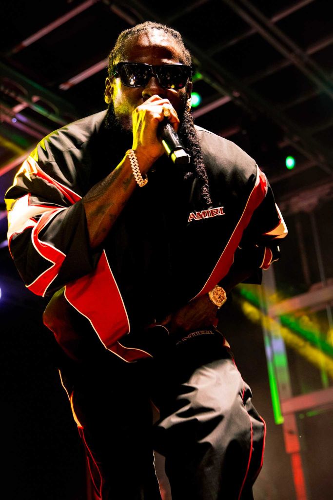 A man wearing sunglasses and a black and red jersey with the word "AMIRI" performing on stage.