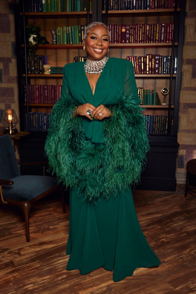 A woman in a green feathered dress and statement necklace stands in front of a bookshelf, smiling confidently.