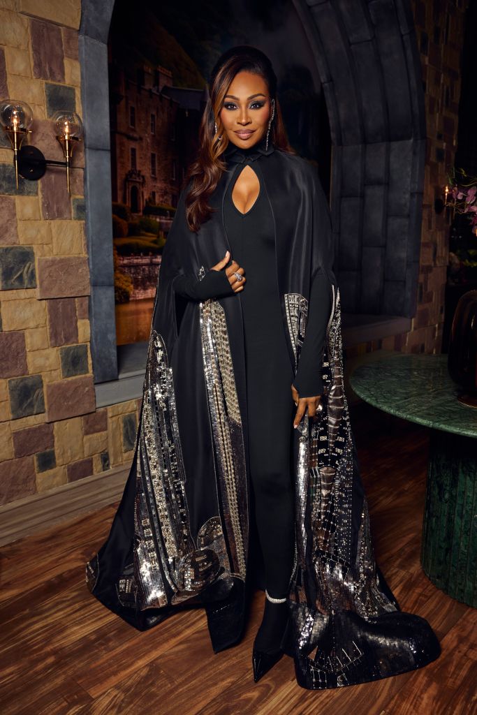 A woman wearing a black dress with sequined sleeves and a high collar stands in a dimly lit stone archway, looking directly at the camera.