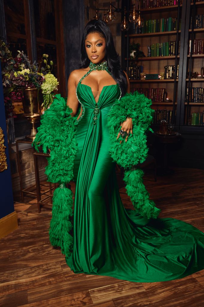 A woman wearing a green feathered dress with a plunging neckline and long train, standing in a room with bookshelves and decorative items.