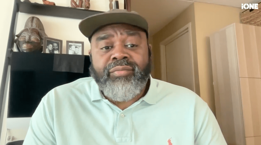A middle-aged Black man with a beard wearing a cap and white shirt, looking directly at the camera with a serious expression.