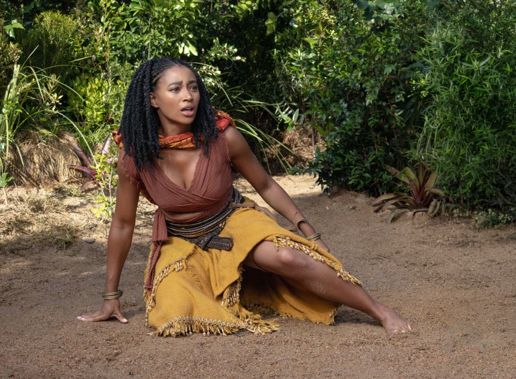 A person with long dark hair wearing a brown top and yellow skirt, sitting on a dirt path in a lush, green environment.